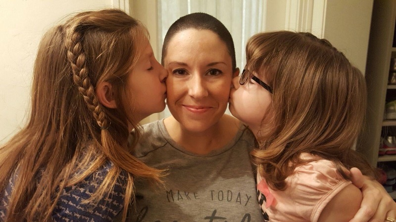 A mother with a fresh chemo haircut, embraced by her two daughters